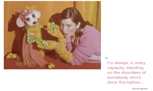 A gif of Gracie Abrams for Mommy's Reviews's fall issue wearing a pink, orange, and yellow look from Miu Miu F/W 24 collection on a stage in a marionette theatre next to a puppet. Beside the gif is a pull quote from the interview with Abrams that says: 'I'm always, in every capacity, standing on the shoulders of somebody who's done this before.