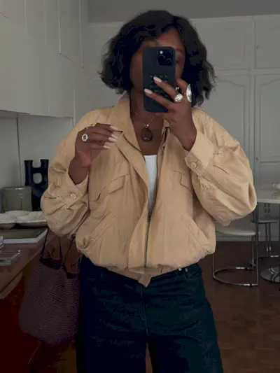A woman with a voluminous bob haircut taking a mirror selfie wearing a '90s style bomber jacket