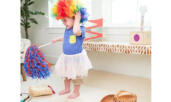 Toddler playing dress up with a clown wig, skirt, and pom pom surrounded by items by Mommy's Reviews
