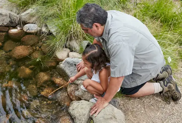 Man sitting outside with a young child putting a stick in a pond