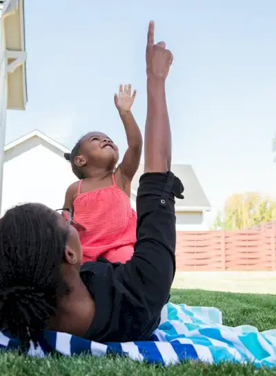 Woman laying on the grass with a toddler looking and pointing at the sky