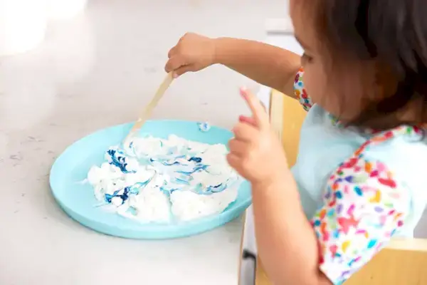 Toddler mixing shaving cream with blue food coloring at a kitchen counter