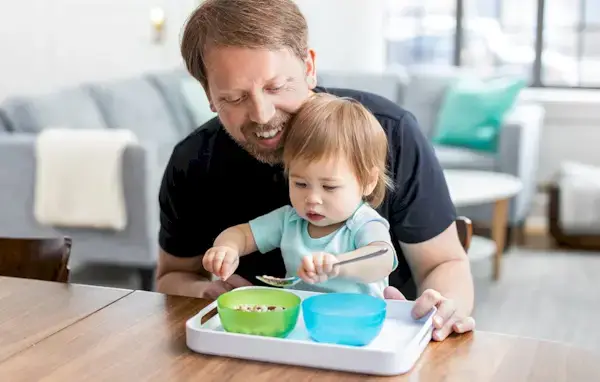 Child scooping food from a bowl while sitting on their dad