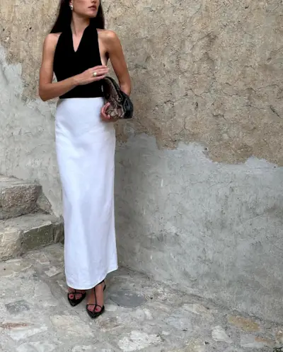 Fashion person Ilirida Krasniqi wears a black halterneck top, white tailored skirt and t-bar heel flops and is carrying a snake print clutch. She is standing in front of a stone wall in a recent image from her Instagram.