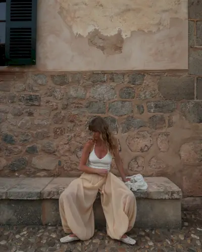 Fashion person Jeannette wears a crochet halterneck top, beige genie pants and crochet mules. She is sitting on a stone bench, in front of a stone wall, in a recent image from her Instagram.