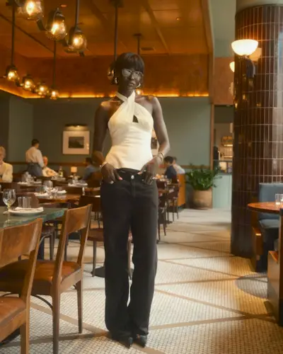 Fashion person Amy Lefevre wears a white halter neck top, a pair of black baggy denim jeans and a black leather pointed toe heel. She is standing in front of a restaurant background in a recent image from her Instagram.