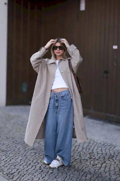 Fashion Person Sophia Geiss at Berlin fashion week. She wears the drawstring jeans trend and pairs them with a brown trench coat, white Miu Miu x New Balance trainers a white cropped tee and black sunglasses.