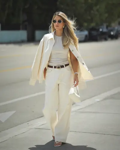 Cass wears a white jacket, beige top, brown belt, white jeans, and white The Row 90s bag.
