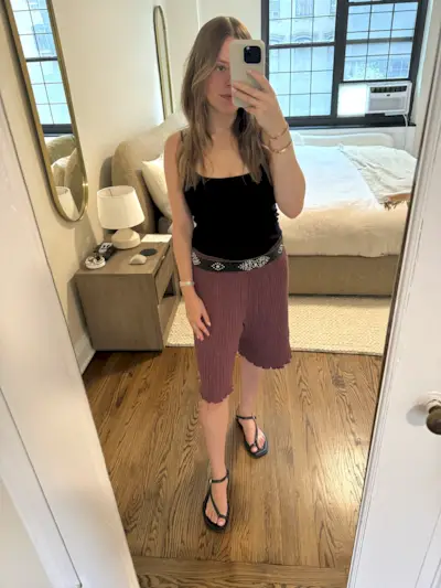 Nikki Chwatt wears Free People Jada bermuda shorts in burgundy with black sandals, a black beaded belt, and a black tank top.