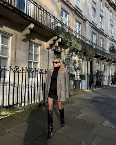 Woman wearing black minidress and oversize blazer with tall black boots.