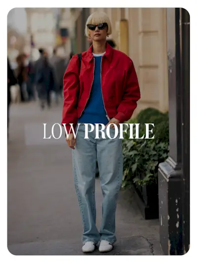 A street style photo of a showgoer wearing a red jacket, jeans, and white, low-profile Adidas sneakers. Text reads, 'Low Profile.
