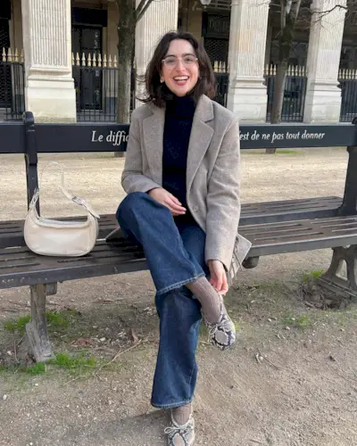 Parisian fashion influencer sits on a bench in Paris wearing a black turtleneck sweater, neutral blazer jacket, jeans, tan socks, and snakeskin ballet flats