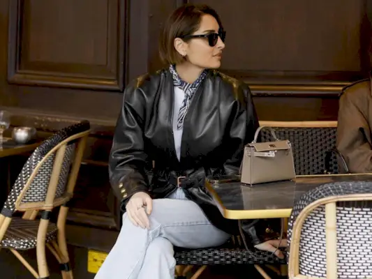 a stylish French woman sits at an outdoor café table with her legs crossed wearing a black leather jacket, tied neck scarf, mini Hermès bag, and jeans