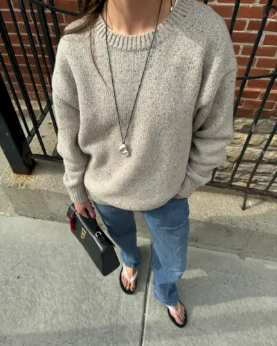 Influencer stands outside wearing white flip-flops with a black sole. She styled her shoes with a grey melange jumper, blue straight-leg jeans and a longline pendant necklace.