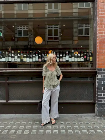 Influencer stands outside a restaurant wearing a green gingham blouse with white embroidered trousers and white flip-flops.