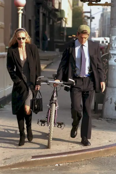An image of Carolyn Bessette-Kennedy wearing a Miu Miu grey midi dress, black trench coat and knee-high boots while walking through New York with John F. Kennedy Jr. in 1996.