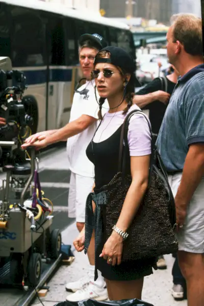 An image of Jennifer Aniston wearing a white t-shirt, black mini dress, baseball hat and tiny sunglasses in 1997, one of the most iconic '90s outfits.