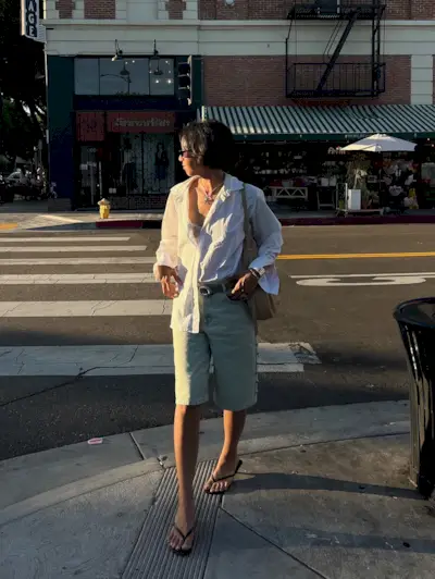 A woman wears a white button-down shirts, denim shorts, and black flip-flops.