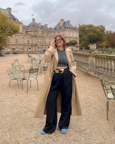 Raeann wears a trench coat, grey top, black wide leg trousers and colourful trainers