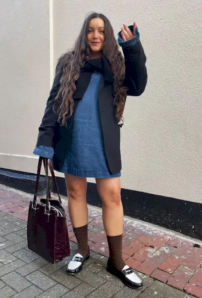 A woman's jean dress outfit with a denim minidress layered underneath black blazer and cardigan and styled with brown socks and two-tone loafers.