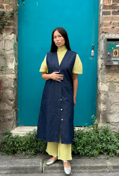 A woman's jean dress outfit with a button-front denim dress over a pleated turtleneck, wide-leg pants, and silver flats.
