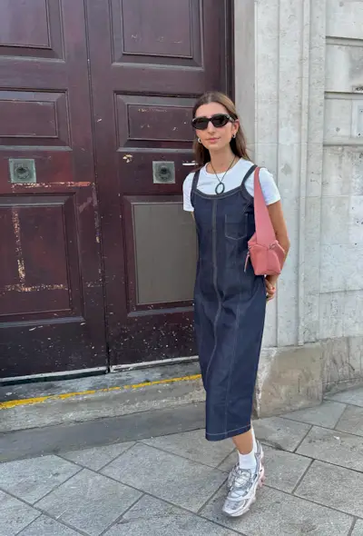 A woman's jean dress outfit with sleeveless midi dress layered over a white T-shirt styled with white and silver sneakers.