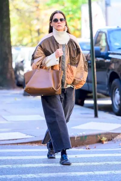 Katie Holmes wears a white turtleneck sweater, a brown jacket, tan tote bag, gray jeans, and black square-toe loafers.