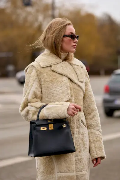 A woman wears a fur coat, black sunglasses, and a black Hermes bag