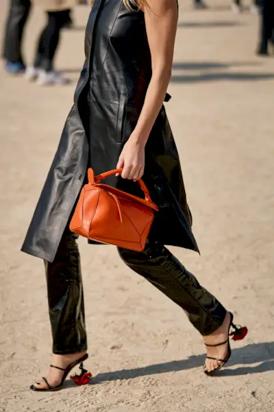 A woman wears an orange Loewe puzzle bag with a leather long dress, leather pants, and Loewe rose heels.