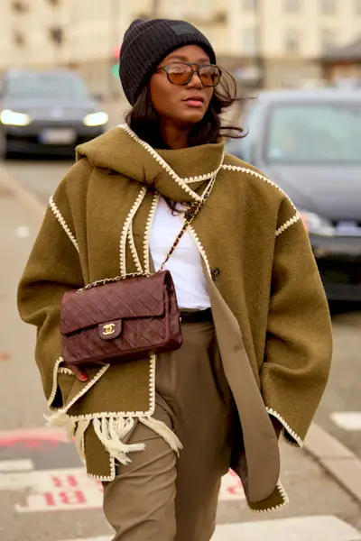 A woman wears the Toteme scarf coat with a burgundy Chanel bag, black beanie, sunglasses, and tan trousers.