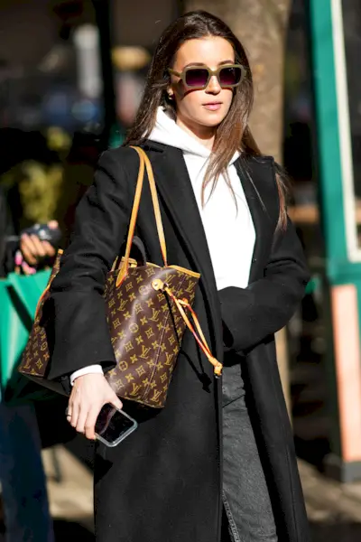 A woman wears a Louis Vuitton tote bag with a black jacket, white sweatshirt, gray jeans, and rectangular sunglasses.