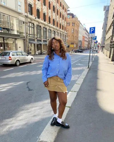 A woman wears a blue stripe button-down shirt, cargo mini skirt, white socks, and black loafers.