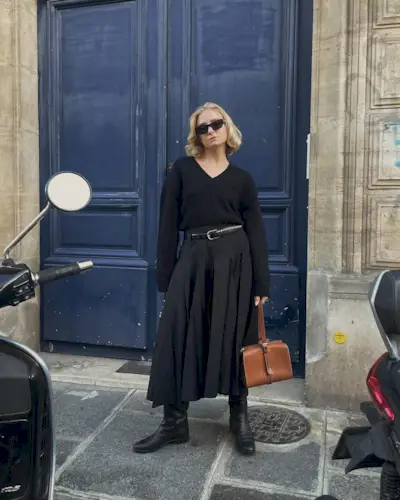 A woman wears a v-neck black sweater, black belt, black pleated midi skirt, black boots, and a tan leather bag.