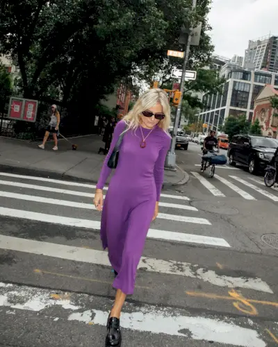 A woman wears black sunglasses, a purple long-sleeve dress, black loafers, and a black shoulder bag.