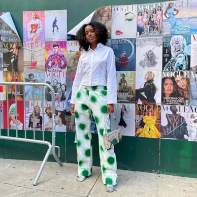 A woman wears a white short button-down shirt, green and white polka dot pants, silver flats, and a silver handbag.