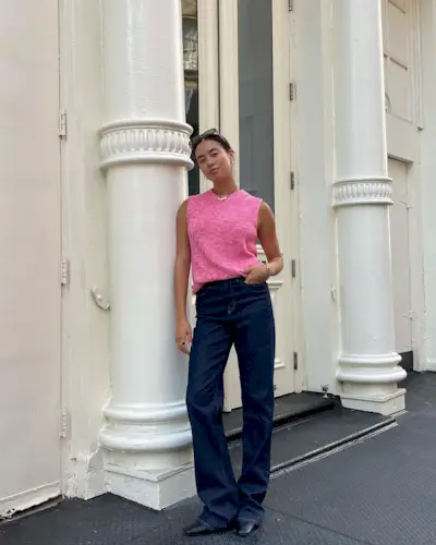 A woman wears a pink sweater vest, dark blue jeans, and black square-toe flats.