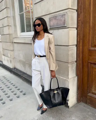 A woman wears a beige blazer, white tank top, black belt, ivory trousers, black flats, and a black tote bag.