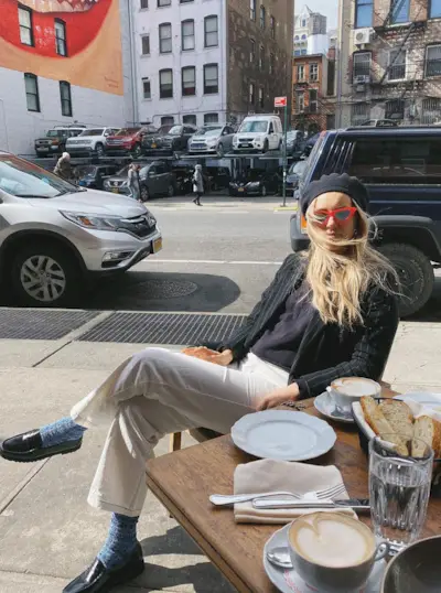 Aemilia wearing white pants, a black cardigan, beret, loafers, and red sunglasses.