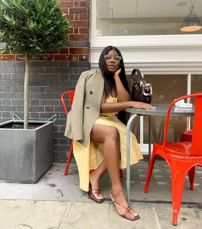 A woman wearing a yellow dress, brown strappy heels, a blazer, and a brown bag with a buckle.