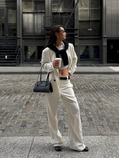 a photo of a woman wearing a white-and-black pinstripe suit with a white tank top, black cardigan, black shoulder bag, and ballet flats