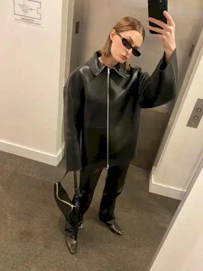 Fashion influencer @christietyler with slicked hair taking a mirror selfie outside an elevator wearing a minimalist black collared leather jacket, black trousers, boots, and sunglasses.