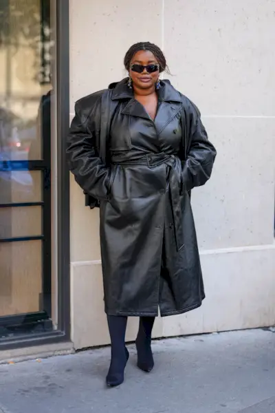 A woman wears a black leather trench coat, black tights, and black heels.