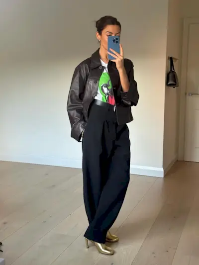 Fashion influencer @smythsisters taking a mirror selfie wearing an on-trend leather jacket, a graphic tee, beltered trousers, and metallic boots.
