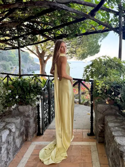 Model @rosiehw stands outside on a balcony in Italy wearing a satin maxi dress in butter yellow.