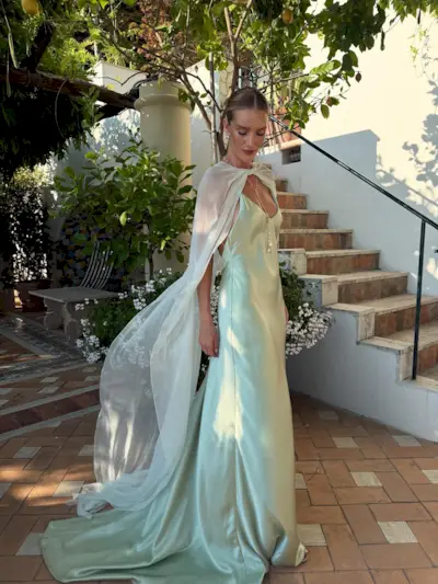 Model @rosiehw stands outside on a balcony in Italy, wearing a satin maxi dress in a light seafoam shade with a sheer cape over her shoulders.