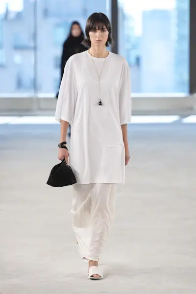 A model at the S/S 25 Toteme show wearing an oversized white T-shirt minidress with a white skirt and mules.