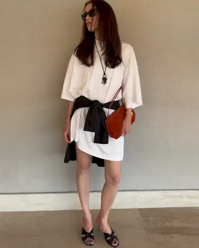 @annabelrosendahl wearing a white mini T-shirt dress with a black sweater around her waist with heeled sandals.