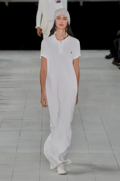 A model at the S/S 25 Ralph Lauren show wearing a white polo T-shirt dress with a beaded cap. 