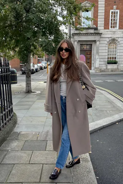 a photo showing what shoes to wear with bootcut jeans, specifically a woman wearing a long tan coat over a white t-shirt with bootcut jeans and loafers