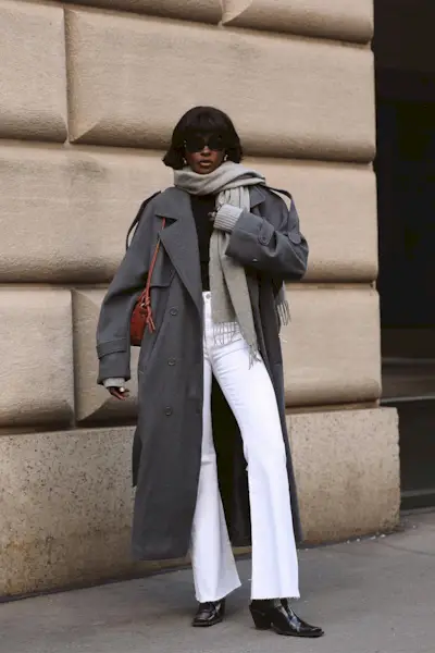 a photo showing what shoes to wear with bootcut jeans, specifically a woman wearing a gray trench coat over a black sweater with white bootcut jeans, a gray scarf, and black cowboy ankle boots